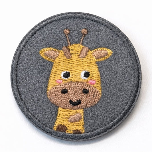 Runder bestickter Giraffen-Aufnäher mit goldgelbem Giraffenkopf, braunen Flecken, großen Augen, rosa Wangen und zwei Hörnchen auf grauem Grund. Ø 5cm, zum Aufbügeln oder Aufnähen.