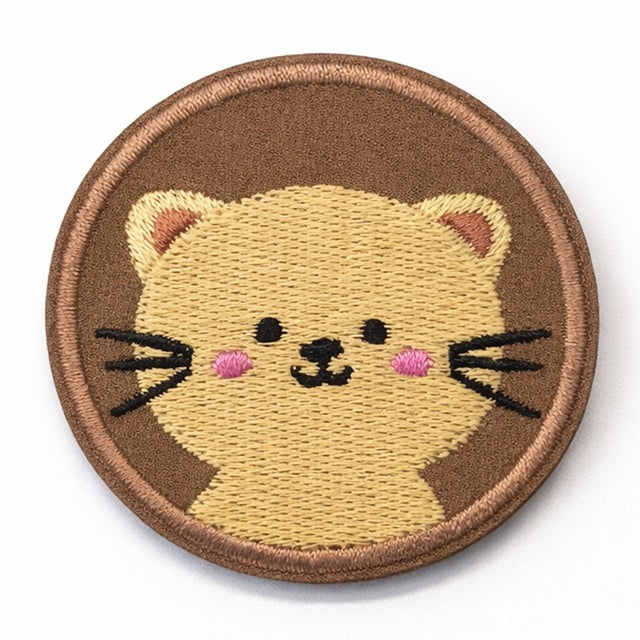 Runder bestickter Katzen-Aufnäher mit cremefarbenem Kätzchen-Gesicht, schwarzen Schnurrhaaren, rosa Wangen und rötlichen Ohren auf braunem Grund mit rosé-goldenem Rand. Ø 5cm, zum Aufbügeln oder Aufnähen.